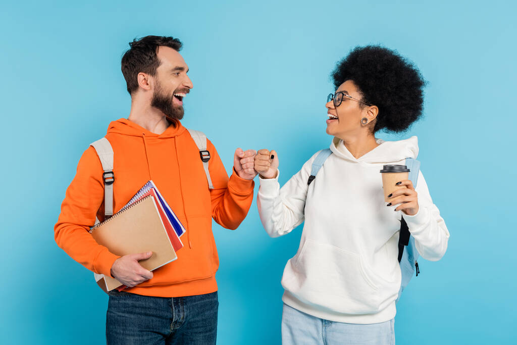 cheerful interracial students with coffee to go and notebooks doing fist bump isolated on blue - Photo, Image