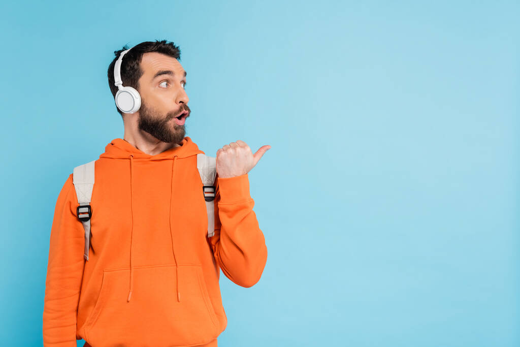 astonished student in wireless headphones and orange hoodie looking away and pointing with thumb isolated on blue - Photo, image