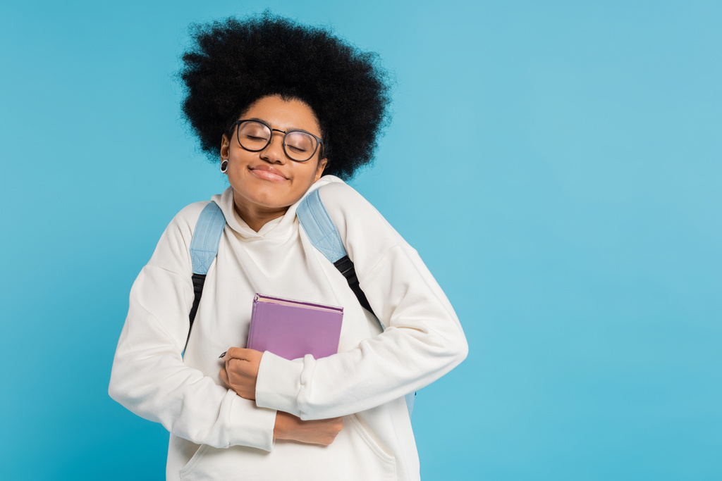 pleased african american student in eyeglasses holding book while smiling with closed eyes isolated on blue - Photo, image