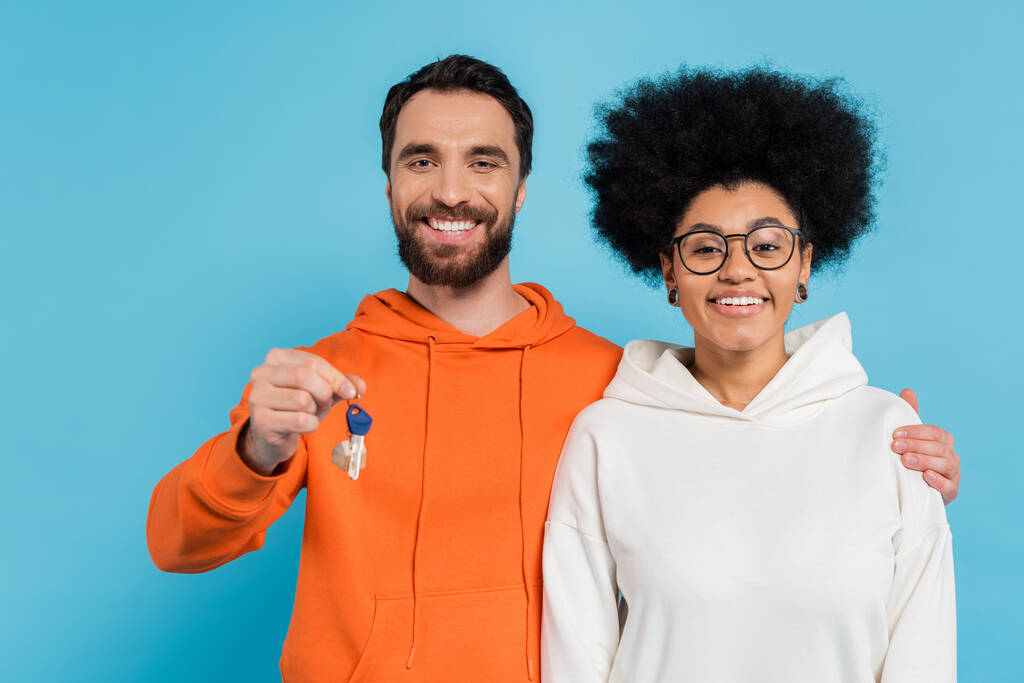 joyful bearded man hugging african american girlfriend and showing key isolated on blue - Photo, Image