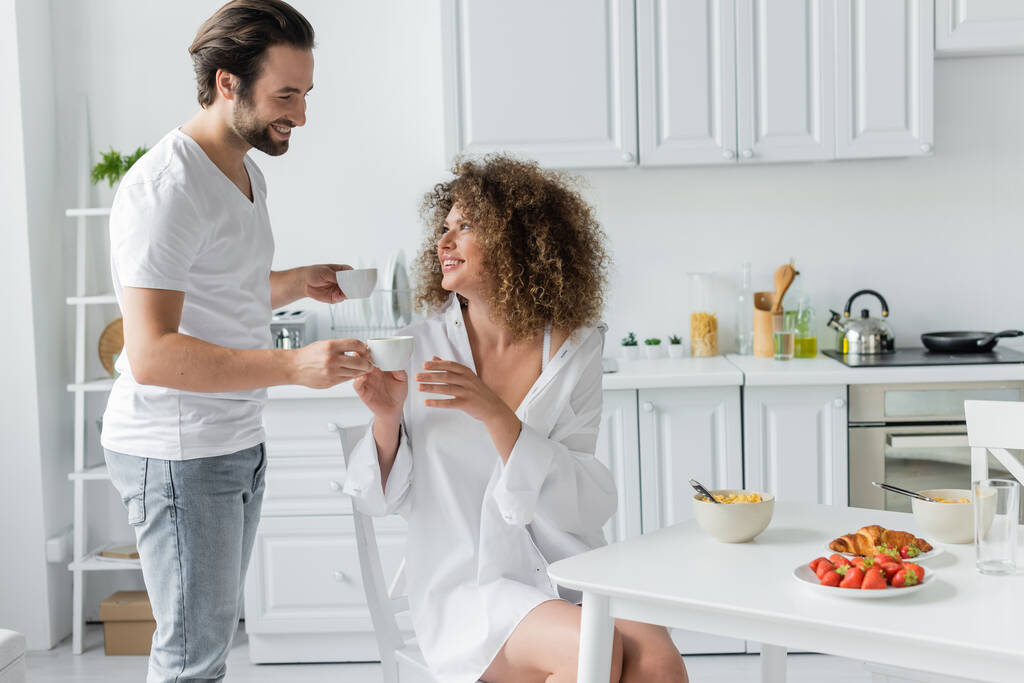 happy young man giving cup of coffee to curly girlfriend during breakfast in kitchen  - Zdjęcie, obraz