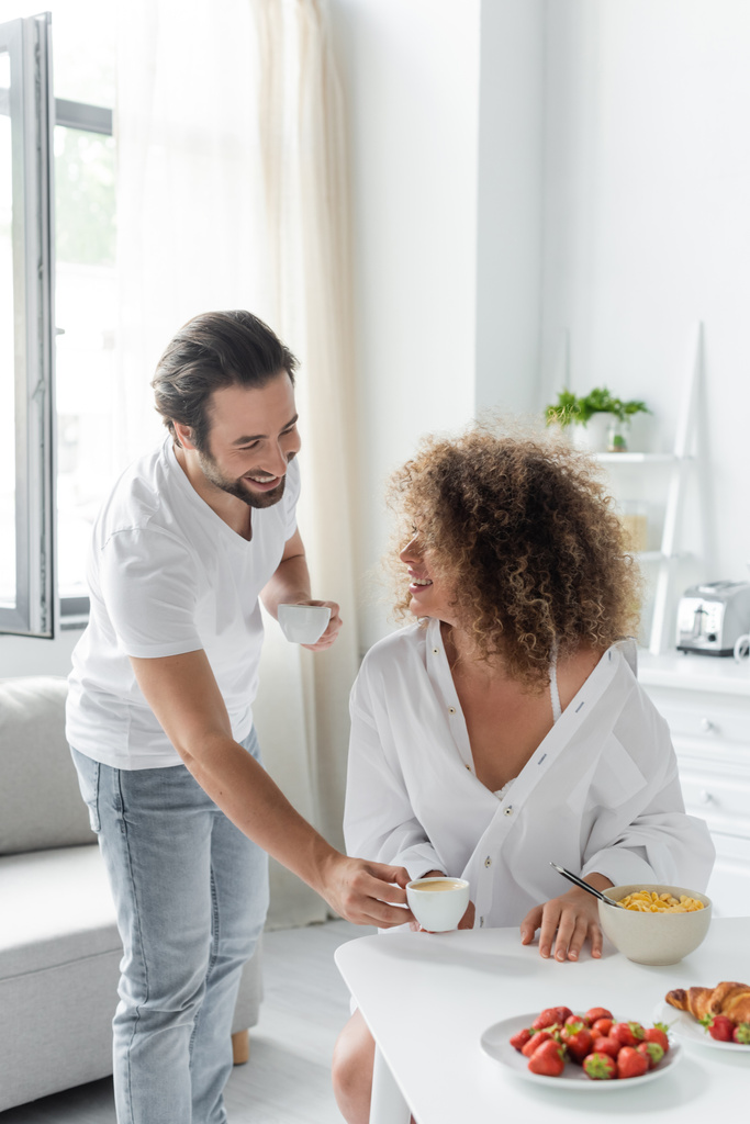 happy man giving cup of coffee to young curly girlfriend during breakfast in kitchen  - Foto, Bild