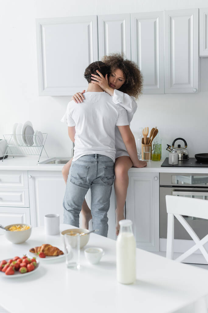 back view of man in jeans seducing curly woman sitting on kitchen worktop  - Foto, imagen