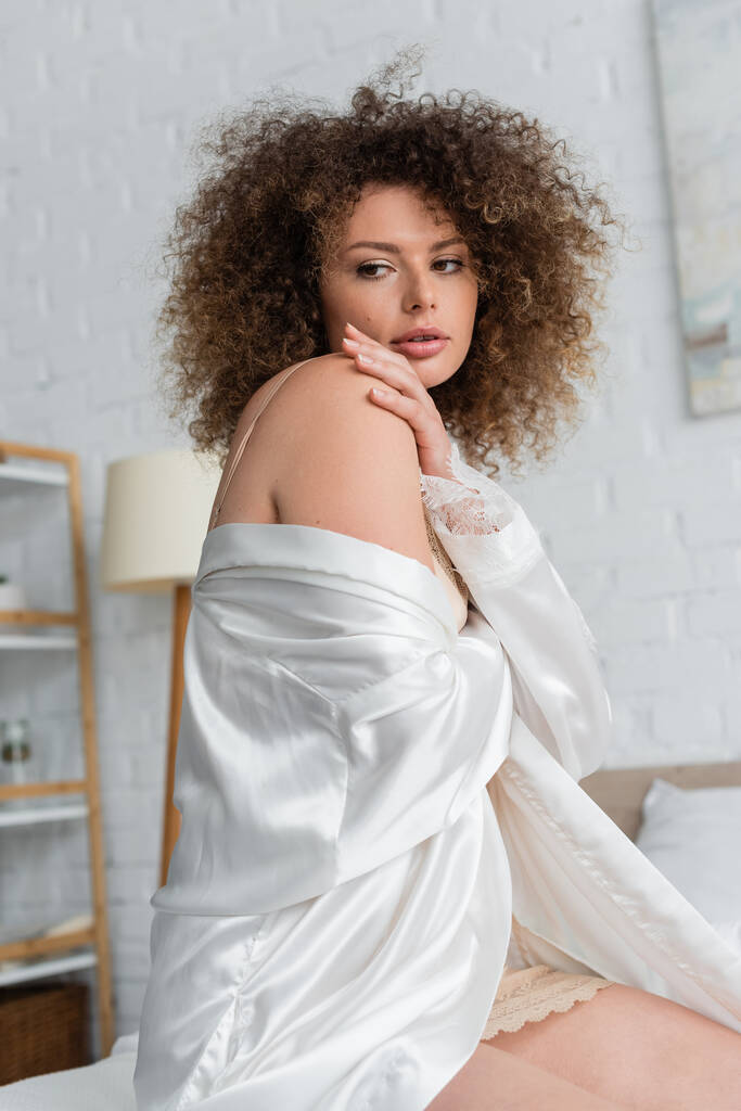 curly and sexy young woman in white silk robe touching shoulder  - Foto, immagini