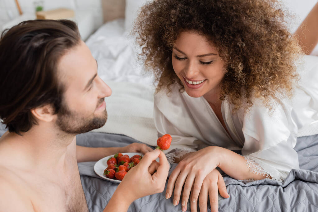 shirtless man holding ripe strawberry near cheerful young woman in silk robe - Foto, Imagen