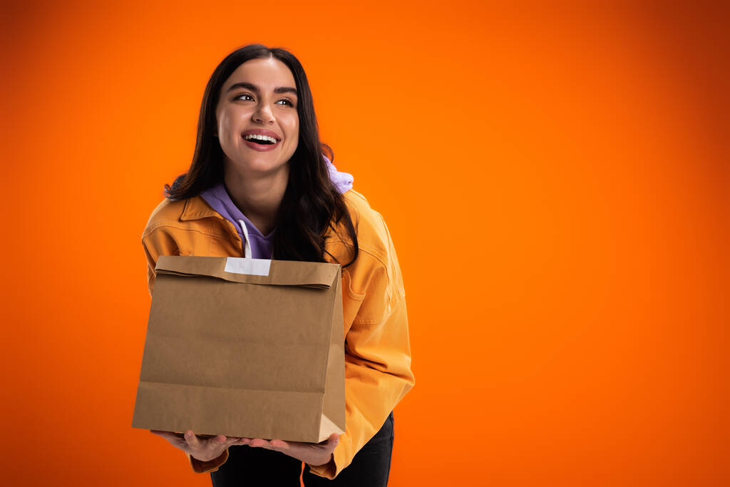 Веселая женщина держит бумажный пакет с едой изолированы на оранжевый - Фото, изображение
