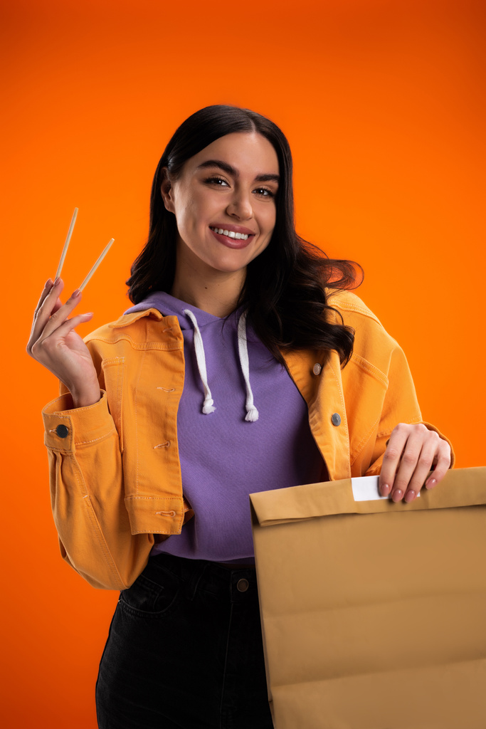 Smiling woman holding chopsticks and paper bag isolated on orange - Foto, imagen