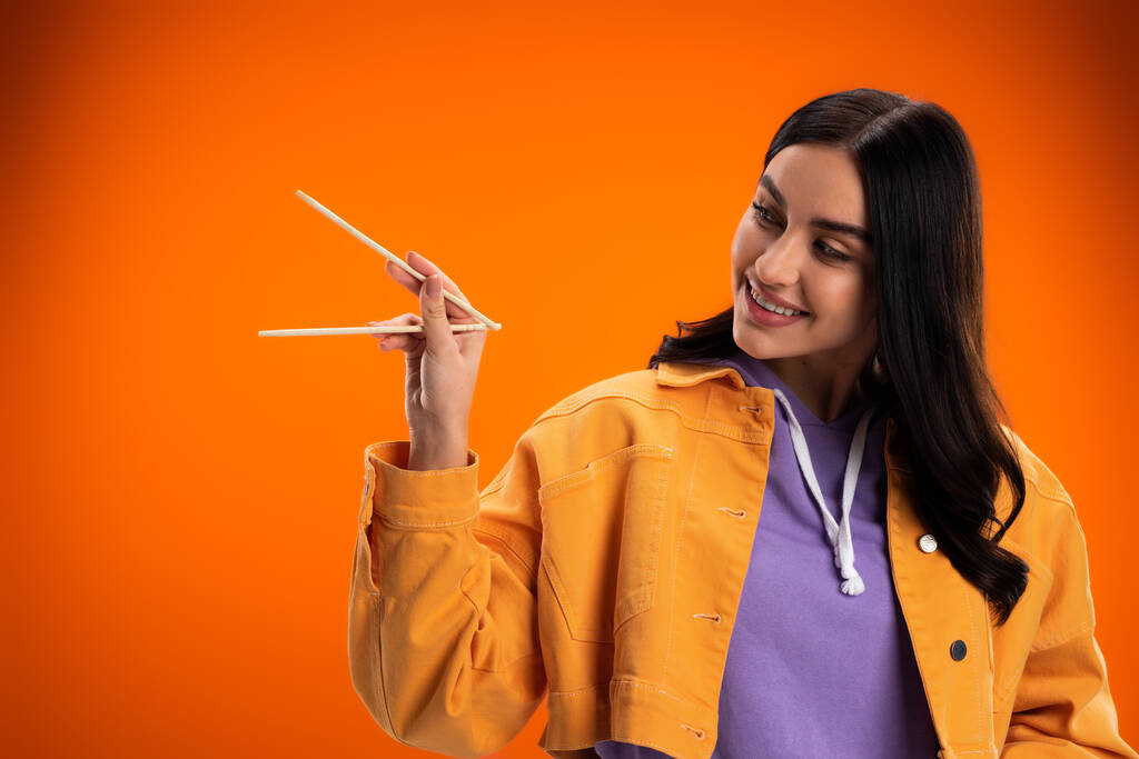 Young brunette woman looking at bamboo chopsticks isolated on orange - Photo, image