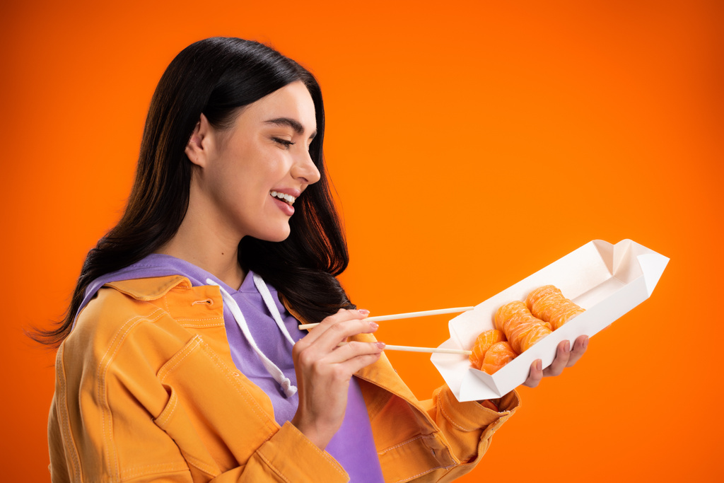 Smiling brunette woman holding takeaway sushi and chopsticks isolated on orange - Fotoğraf, Görsel