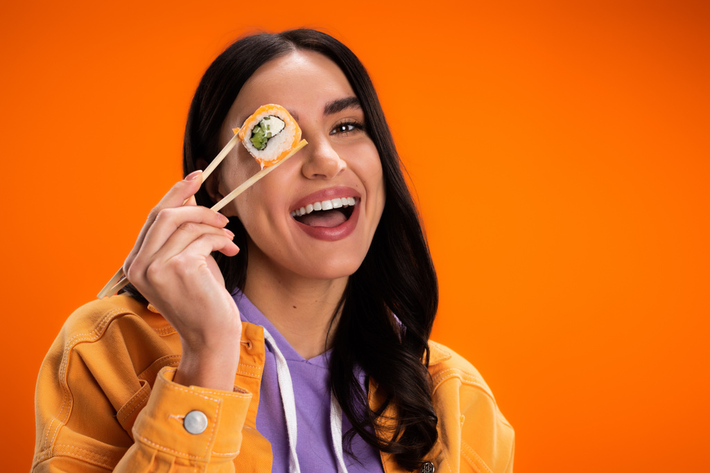 Positive young woman holding sushi in chopsticks near eye isolated on orange - Photo, image