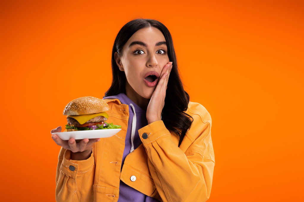 изумленная женщина в модной куртке держит вкусный бургер, трогая лицо и глядя на камеру, изолированную от оранжевого - Фото, изображение