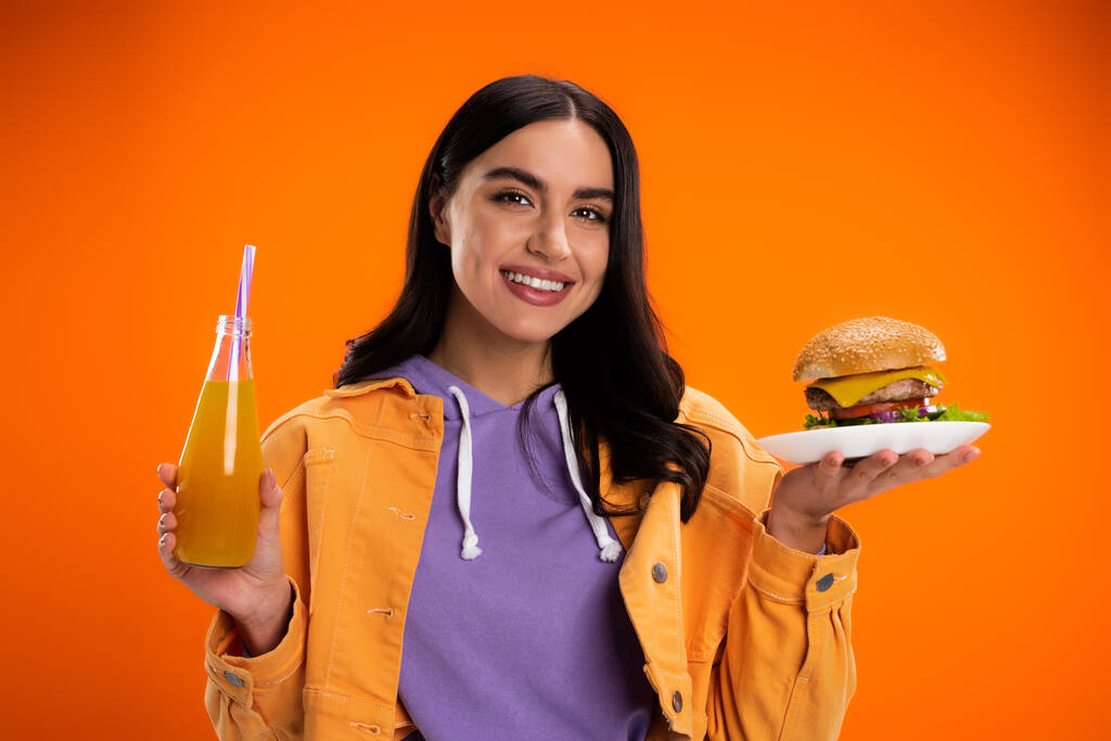 おいしいハンバーガーと新鮮なレモネードのボトルを持つ幸せな女性はオレンジに孤立したカメラを見て - 写真・画像