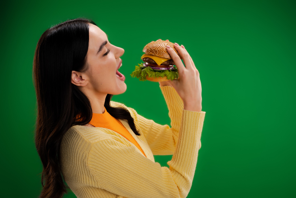 緑に囲まれたチーズと肉でハンバーガーの近くで口を開ける若い空腹の女性の側面図 - 写真・画像