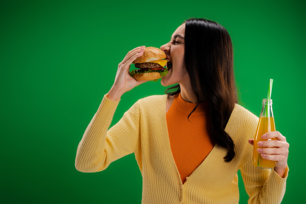 ブルネット女性保持ボトルの新鮮なレモネードと食べるおいしいバーガー隔離された緑 - 写真・画像