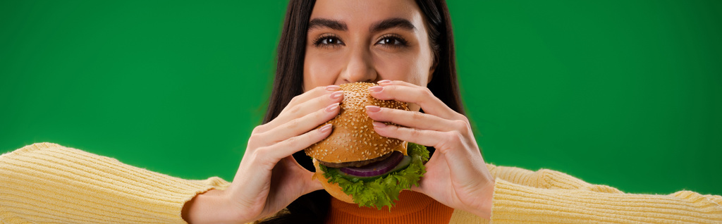 若い空腹の女性がハンバーガーを食べて緑のバナーに隔離されたカメラを見て - 写真・画像