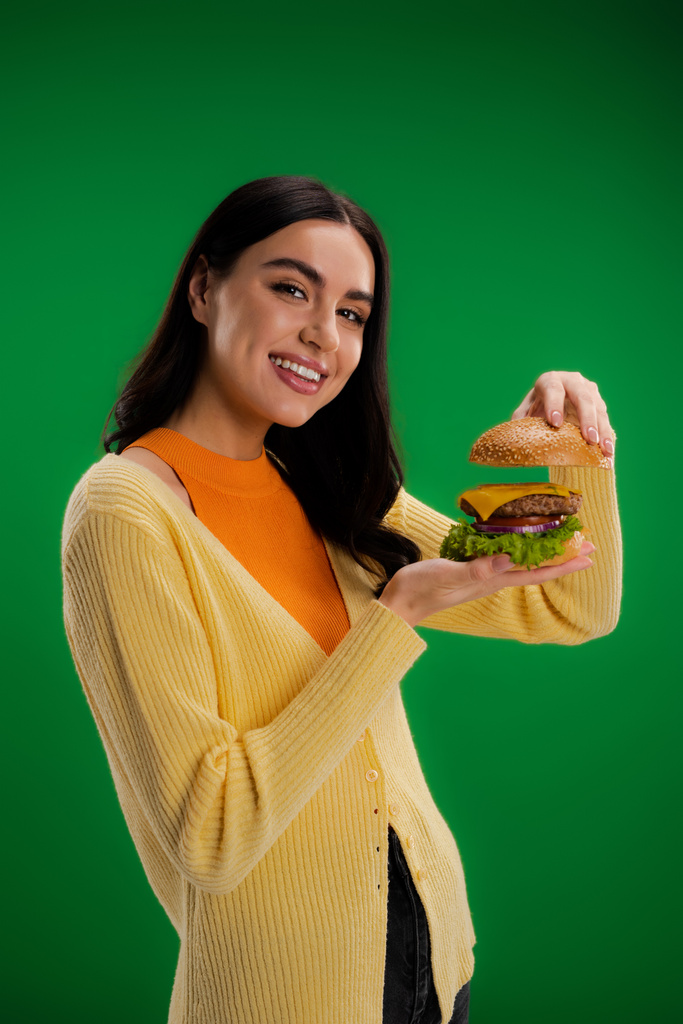 young brunette woman showing delicious burger while smiling at camera isolated on green - Fotó, kép
