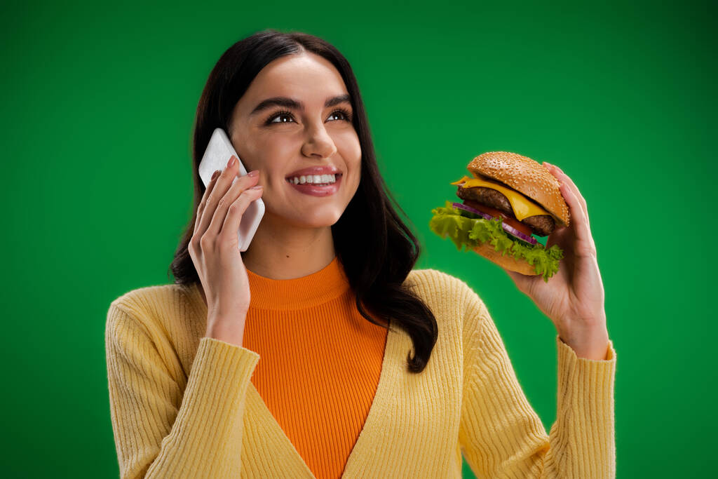happy young woman holding delicious burger and talking on smartphone isolated on green - Fotó, kép