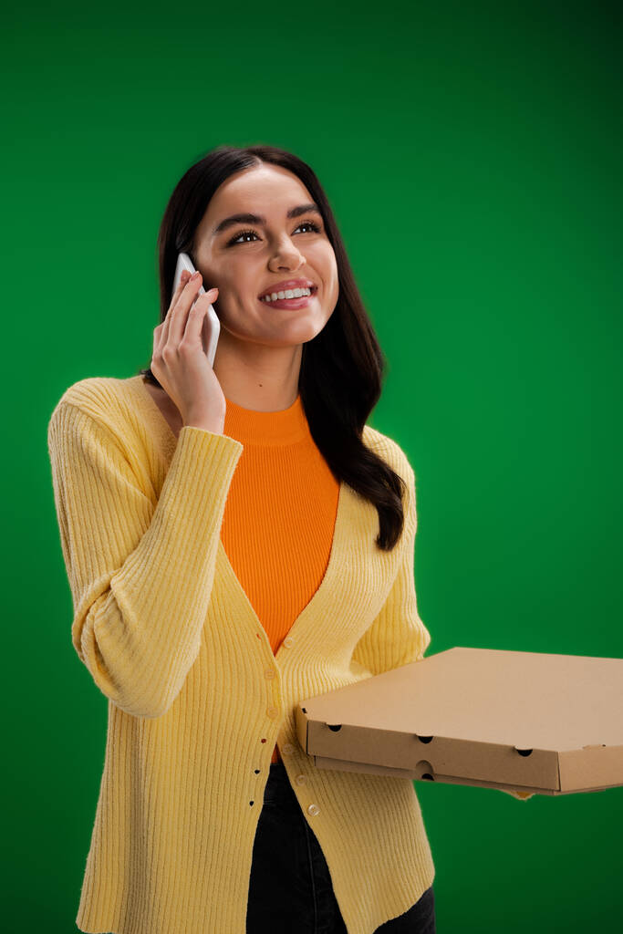 happy young woman calling on smartphone while standing with pizza box isolated on green - Foto, imagen