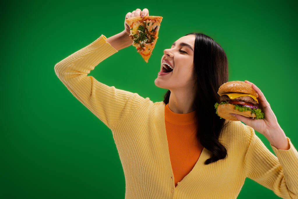 hungry woman holding delicious burger and opening mouth near piece of pizza isolated on green - Fotoğraf, Görsel
