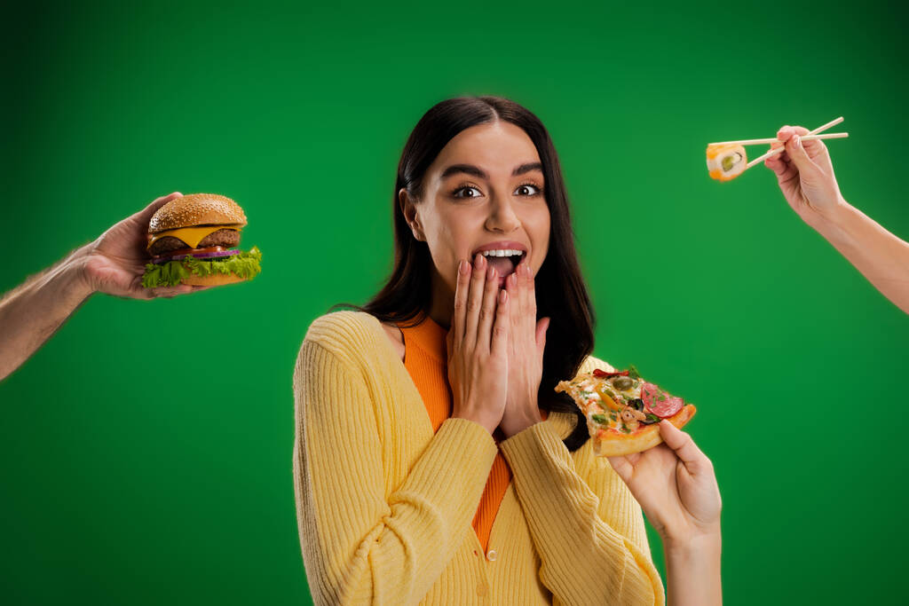 amazed woman covering mouth with hands near people proposing different food isolated on green - Photo, image