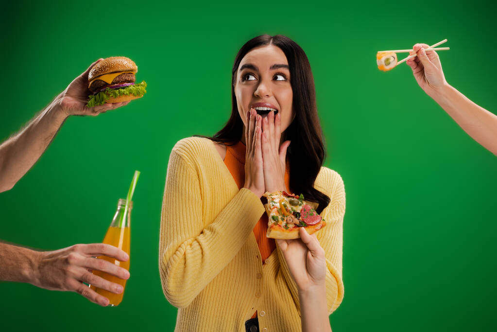 astonished woman covering mouth with hands near people proposing different food isolated on green - Foto, immagini