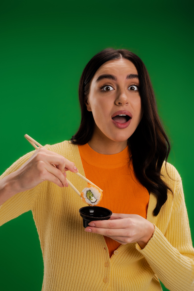 astonished woman holding sauce bowl and chopsticks with sushi roll while looking at camera isolated on green - Photo, image