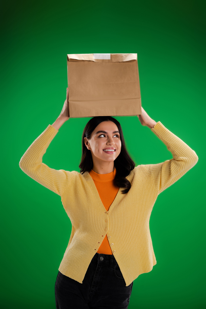 cheerful young woman holding paper bag above head isolated on green - Foto, imagen