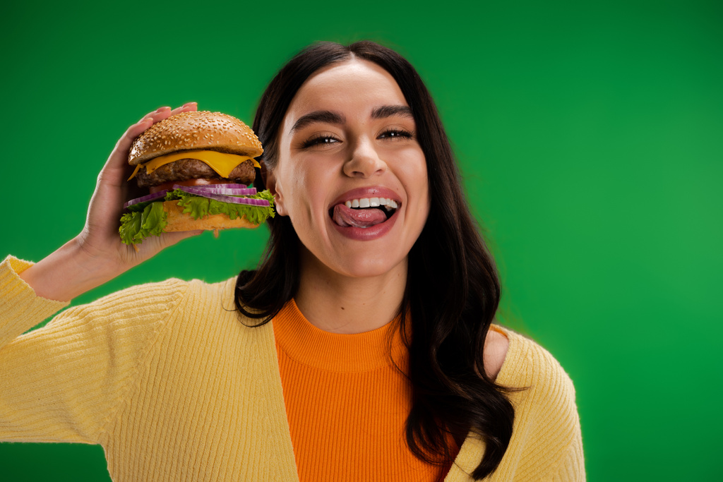 brunette woman holding tasty burger and sticking out tongue isolated on green - Photo, Image
