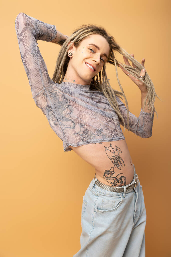 Smiling queer person touching dreadlocks and looking at camera isolated on yellow  - Фото, изображение