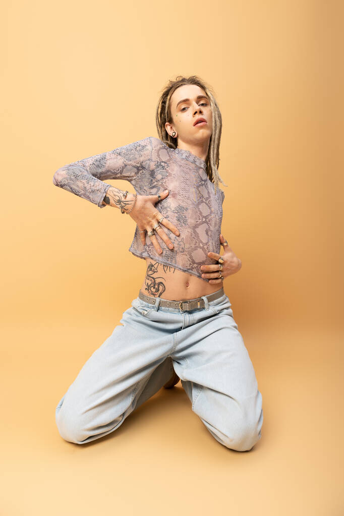 Trendy tattooed queer person touching crop top with snakeskin print on yellow background  - Fotografie, Obrázek