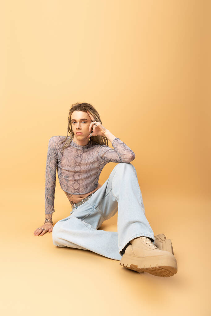 Full length of nonbinary person looking at camera while sitting on yellow background  - Fotografie, Obrázek