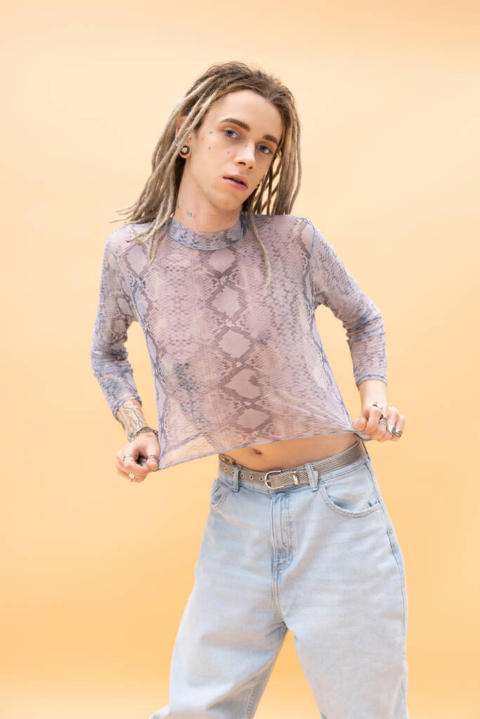 Young queer person with dreadlocks touching crop top with snakeskin print isolated on yellow  - Photo, image