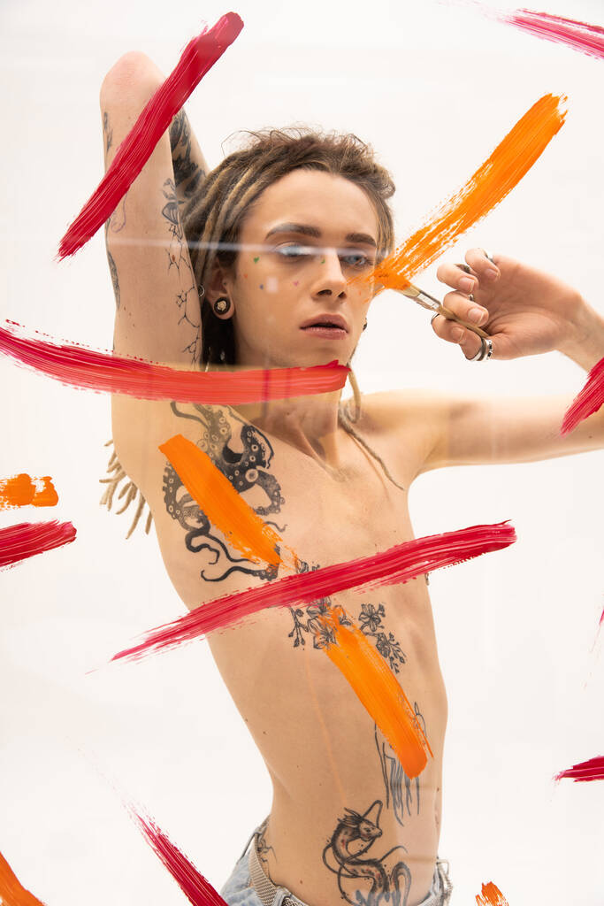 shirtless nonbinary model with tattooed body painting on glass surface while standing on white background  - Foto, afbeelding