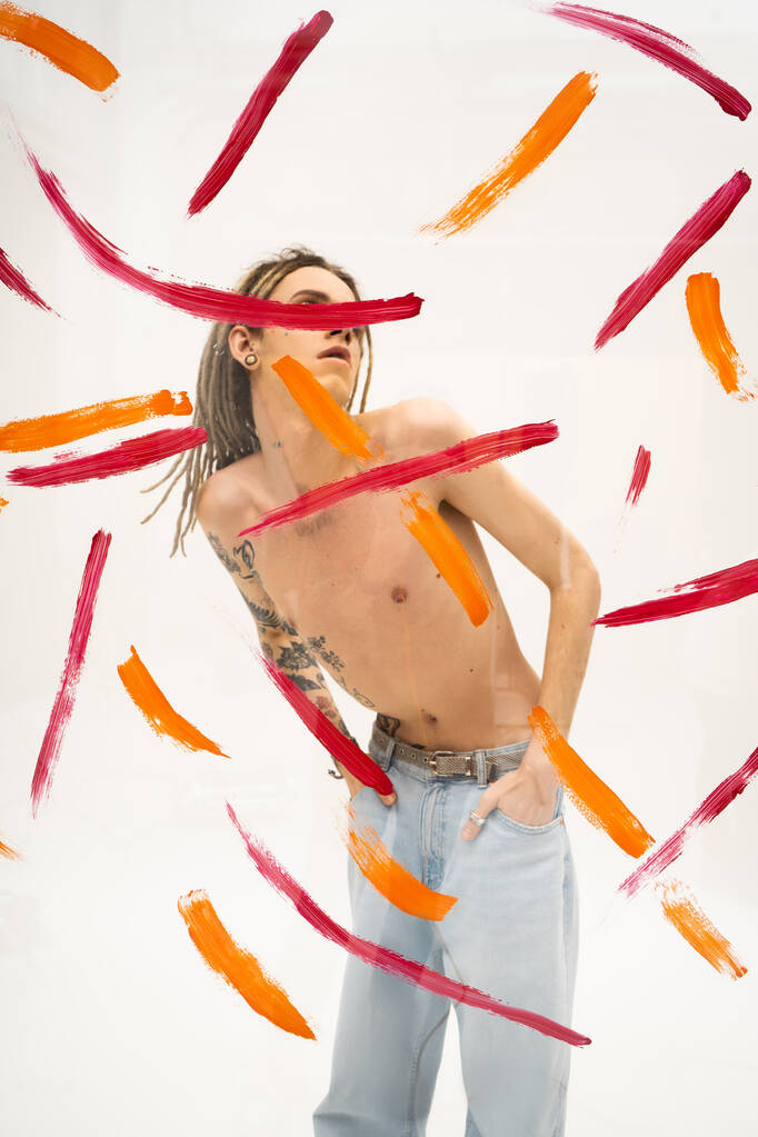 queer person with shirtless tattooed body holding hands in pockets of jeans behind glass with colorful paint strokes on white background - Фото, изображение