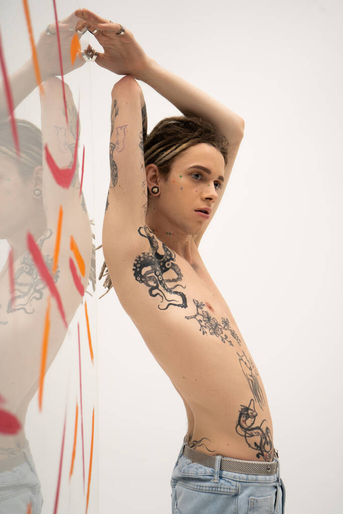 young nonbinary model with tattooed body posing with raised hands near glass with paint spills on white background - Φωτογραφία, εικόνα