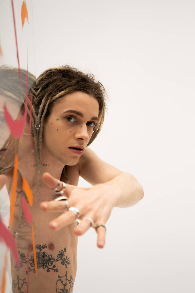 young nonbinary person with silver rings on blurred hand looking at camera near glass with colorful strokes on white  - Foto, imagen