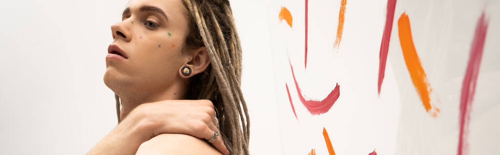 portrait of queer person with dreadlocks posing with hand on shoulder near colorful brush strokes on white, banner - Фото, изображение