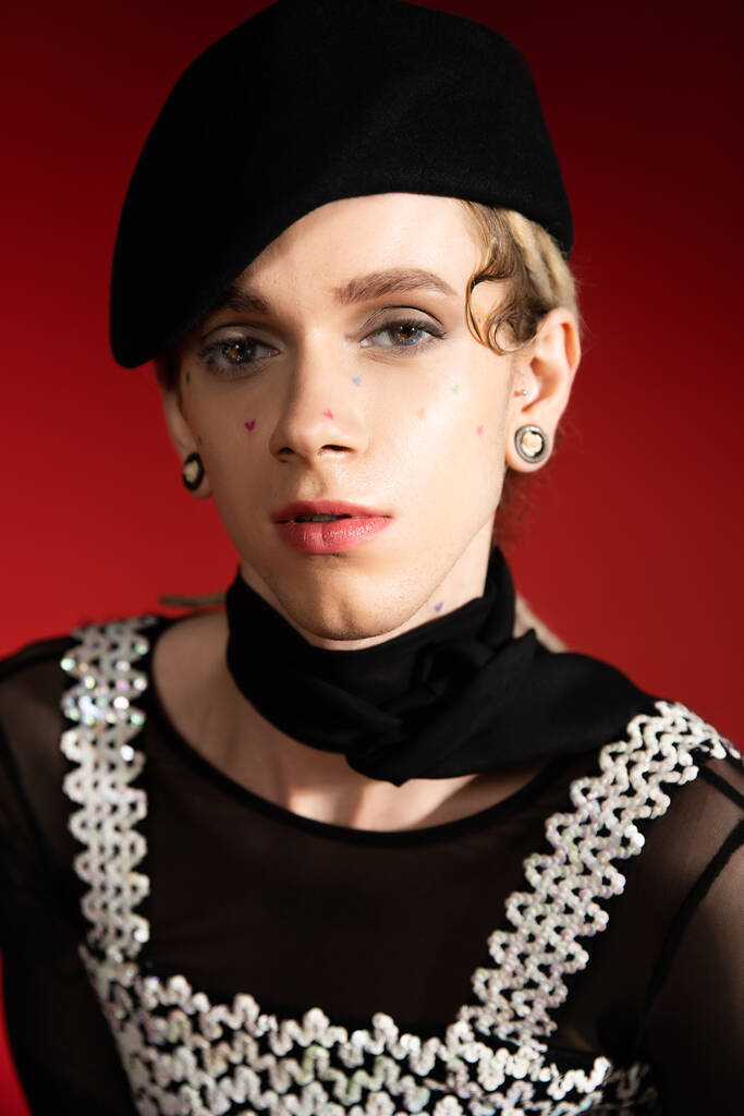 portrait of elegant nonbinary model in black beret and neckerchief looking at camera on red background - Photo, Image