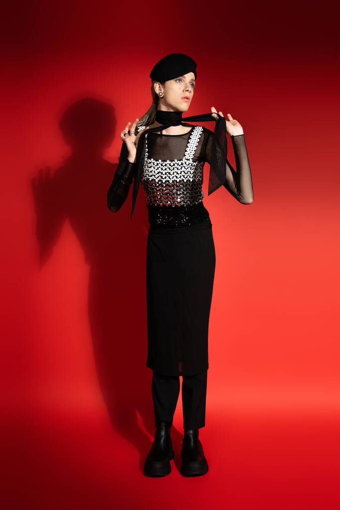 full length of nonbinary model in beret and elegant top tying neckerchief on red background with shadow - Φωτογραφία, εικόνα