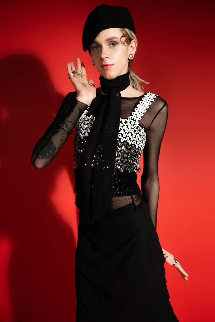 tattooed queer model in elegant attire waving hand and looking at camera on red background with shadow - Fotó, kép