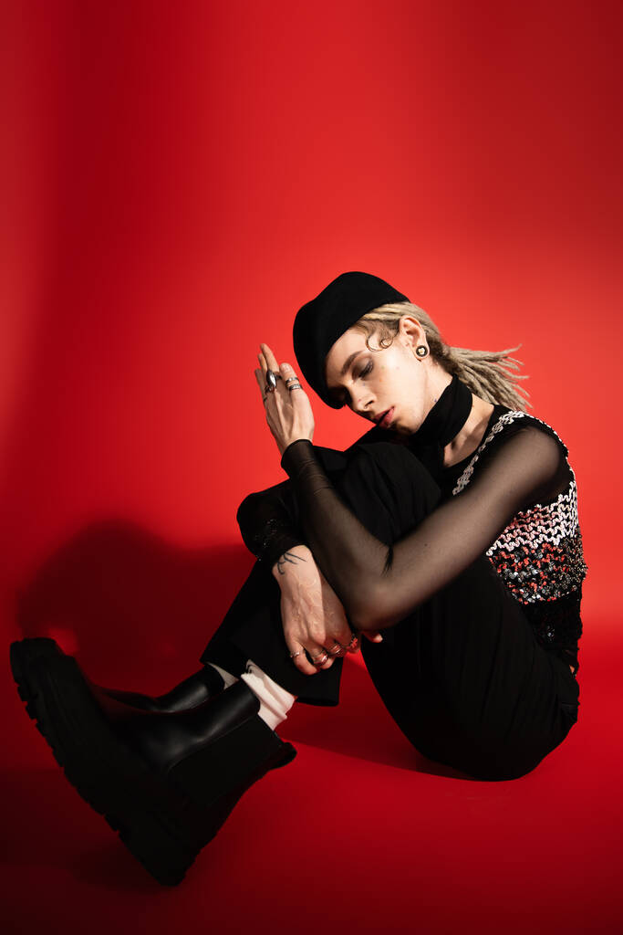 full length of nonbinary model in black boots and beret sitting with closed eyes on red background - Fotografie, Obrázek
