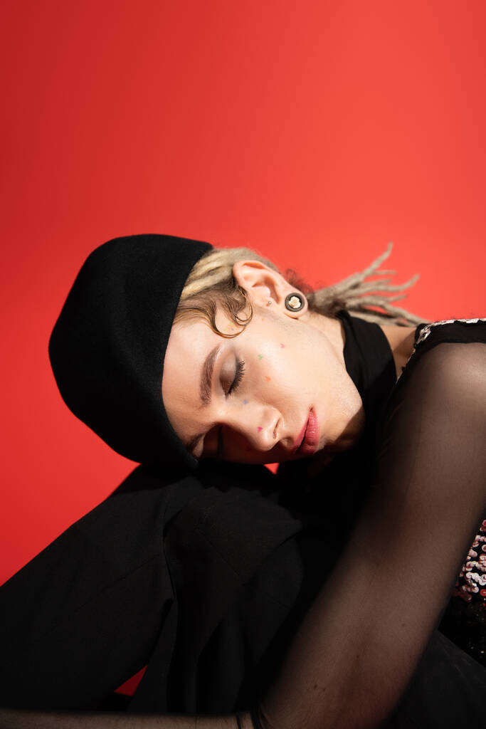 portrait of queer model in black beret sitting with closed eyes isolated on red - Foto, Imagem