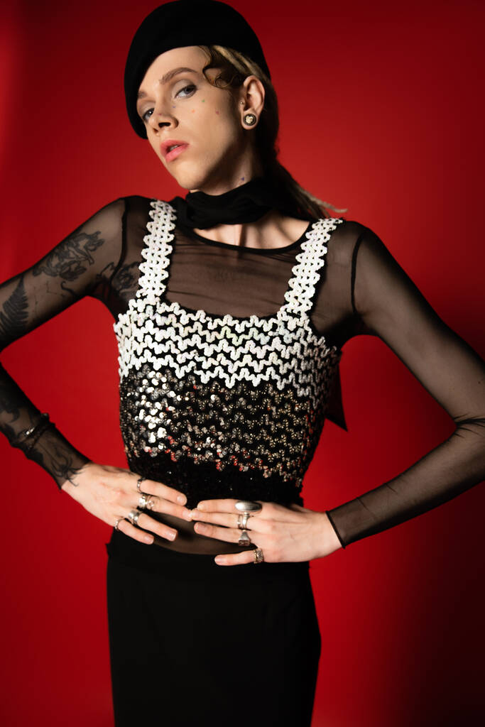 tattooed nonbinary person in elegant clothes and black beret posing with hands on waist on red background - Zdjęcie, obraz