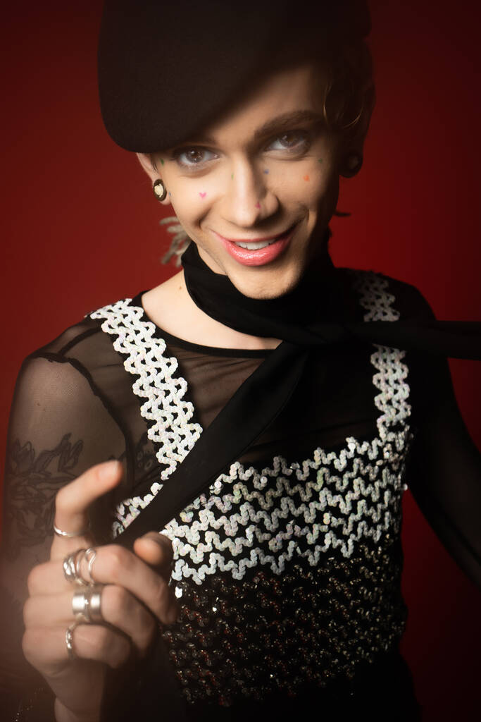 smiling queer model in elegant top and black beret looking at camera and pointing with finger on dark red background - Fotoğraf, Görsel