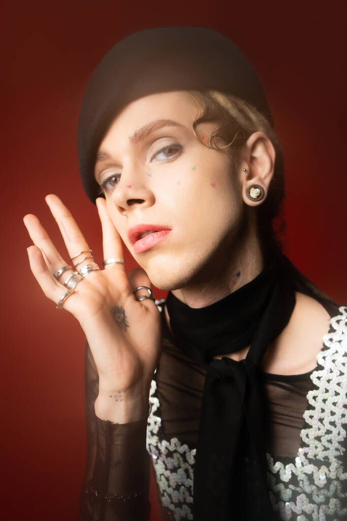 portrait of young queer model in stylish attire and silver rings looking at camera on dark red background - Foto, Imagen