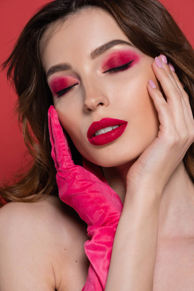 portrait of young woman with magenta color eye shadow gently touching face with hands in glove isolated on pink  - Photo, Image
