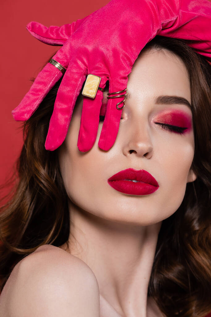 brunette young woman in magenta color glove with golden rings covering eye isolated on pink  - Foto, afbeelding
