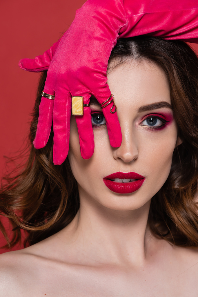 brunette woman in magenta color glove with golden rings covering eye while looking at camera isolated on pink  - Foto, Imagem