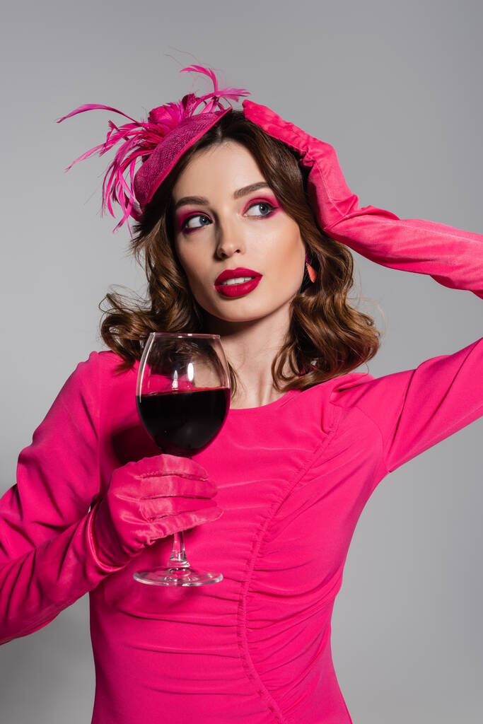 брюнетка жінка в рукавичках кольору пурпуру і елегантний капелюх з пером тримає келих червоного вина ізольовані на сірому
  - Фото, зображення