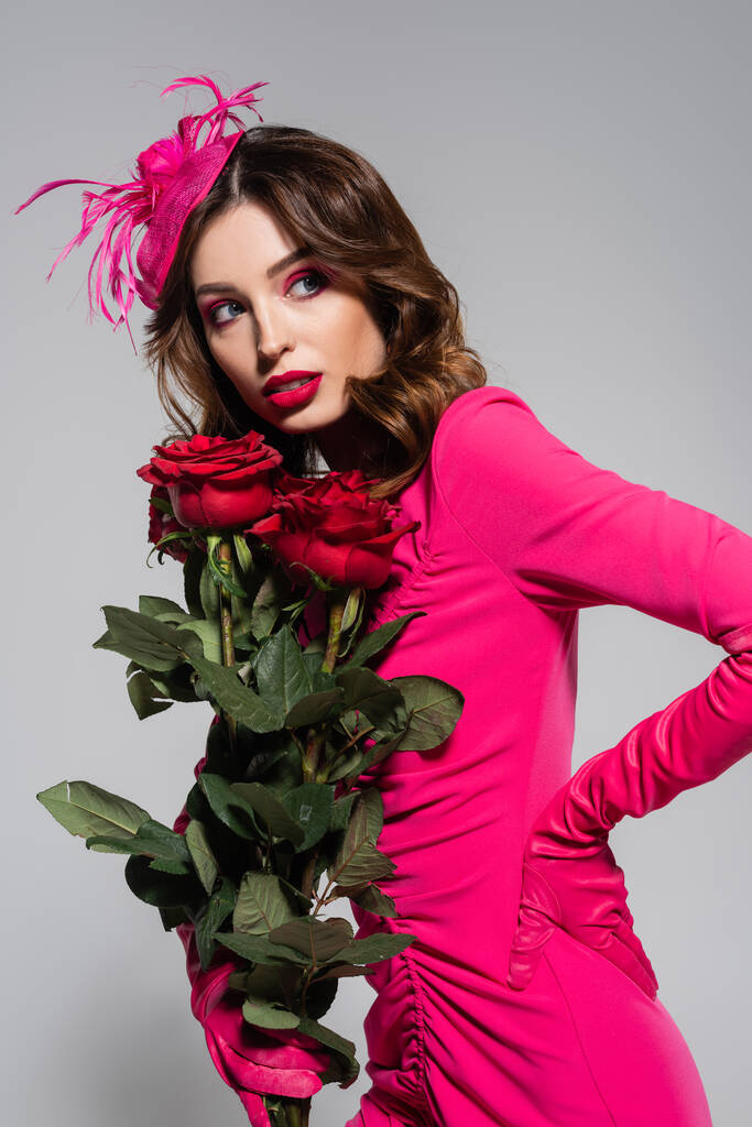 brunette young woman in magenta color gloves and hat posing with hand on hip while holding red roses isolated on grey  - Photo, Image